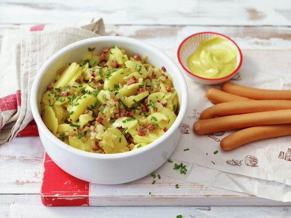 Ein frisch zubereiteter Kartoffelsalat, daneben sind Wiener Würstchen und Senf platziert.