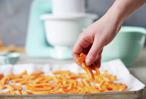 Karotten werden in einer Bosch Küchenmaschine zerkleinert, welche umgeben von Kartoffelsalat und frischem Gemüse ist.
 Bild 2: Süßkartoffelpommes werden sorgfältig auf ein Backblech platziert.