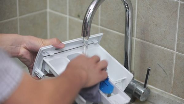 Mani che sciacquano la vaschetta del detersivo della lavatrice sotto il rubinetto