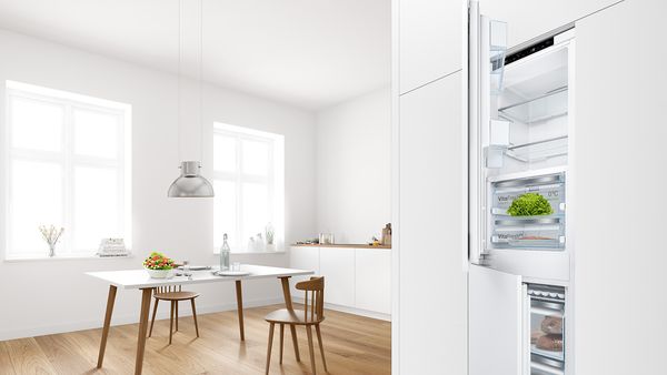 Kalusteeseen sijoitettu Bosch-jääkaappipakastin, jonka ovet ovat hieman raollaan. Vieressä avautuu moderni ruokailuhuone.