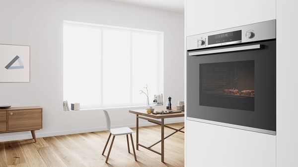 Sort Bosch ovn steger kød i hvidt køkkenskab ved siden af en moderne spisestue i skandinavisk stil.
