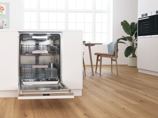Lidt åben Bosch opvaskemaskine indbygget i hvidt køkken. Bordplader og gulve af træ. 