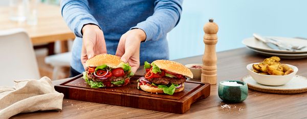 Eine Person richtet zwei Burger, die mit einer Küchenmaschine mit Fleischwolf selbst gemacht wurden, auf einem Holzbrett an.
