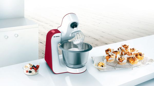 Kleine rotweiße Bosch MUM-Küchenmaschine auf einer weißen Küchentheke, daneben Schokomuffins 
