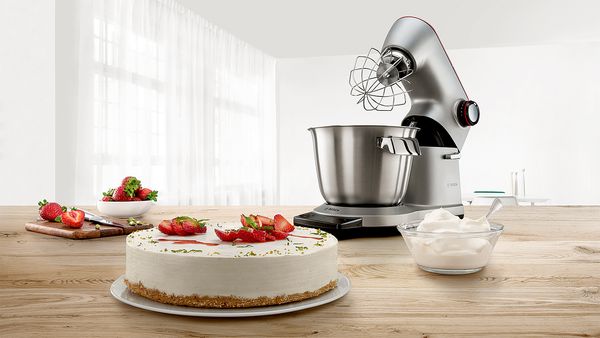 Vor einer silbernen Küchenmaschine von Bosch steht eine Erdbeer-Sahne Torte, aufgeschlagene Sahne und frische Erdbeeren.