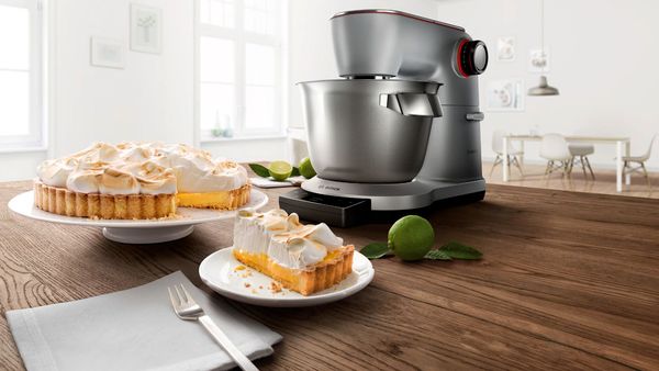 Vor einer silbernen Küchenmaschine von Bosch steht eine frisch gebackene Baiser-Torte, von der ein Stück auf einem Teller neben Besteck und Serviette liegt.