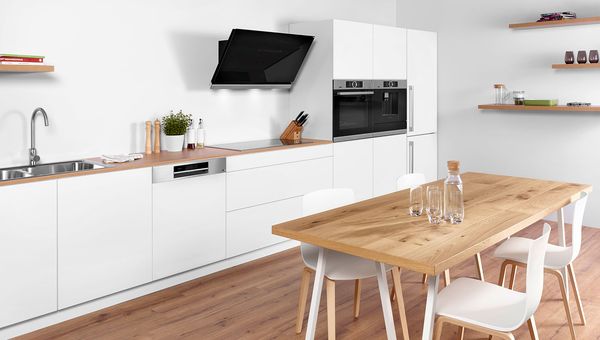White single-wall kitchen in modern design. Wall-mounted cooker hood. Built-in Bosch dishwasher, fridge freezer, oven and fully automated espresso maker. Wood table in foreground with four chairs and water glasses. 