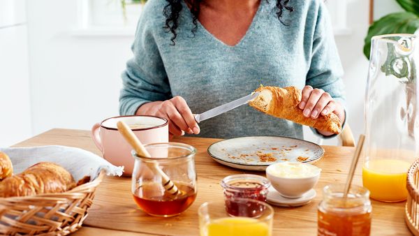 Eine Frau sitzt am gedeckten Frühstückstisch und bestreicht ein Croissant mit Butter.